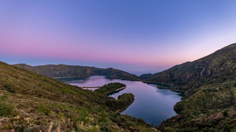 Serene lake at sunset, surrounded by green hills with purple-pink sky.