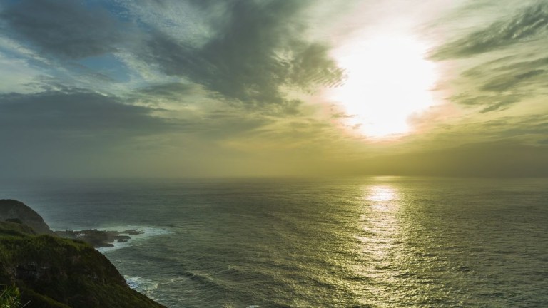 Sunset over the ocean with cloudy sky and coastline on the left.