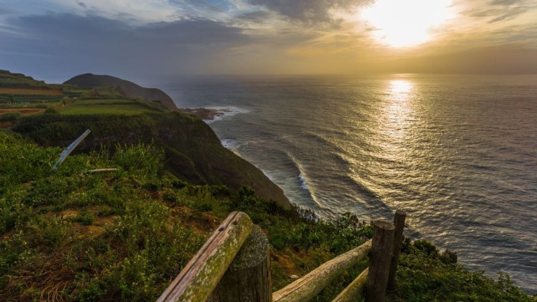 Sunset view over ocean from a grassy cliff with wooden fence.