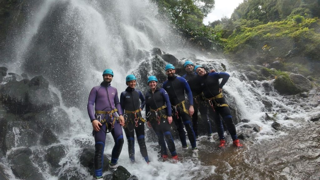Group of people in wetsuits and helmets smiling by a waterfall.