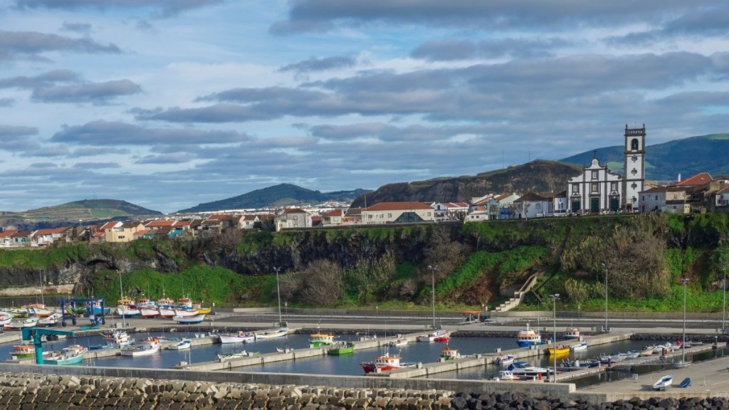 Harbor with boats, green hills, and a distant church under a cloudy sky.