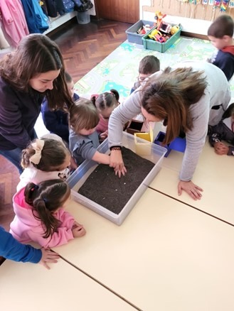 Children and teachers gather around a tray of soil on a classroom table.