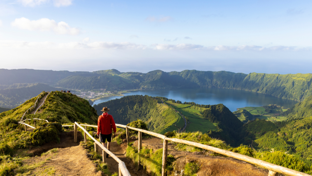 Person in red jacket hiking on a trail overlooking a lake and green mountains.
