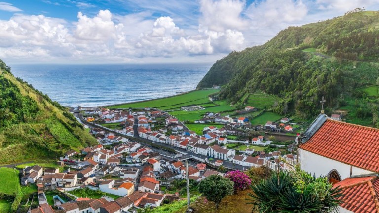 Coastal town with red-roofed houses, green hills, and ocean view under a cloudy sky.