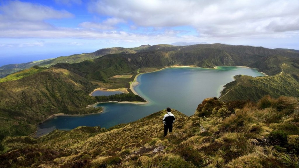 Person standing on a hill overlooking a large lake with mountains under a cloudy sky.
