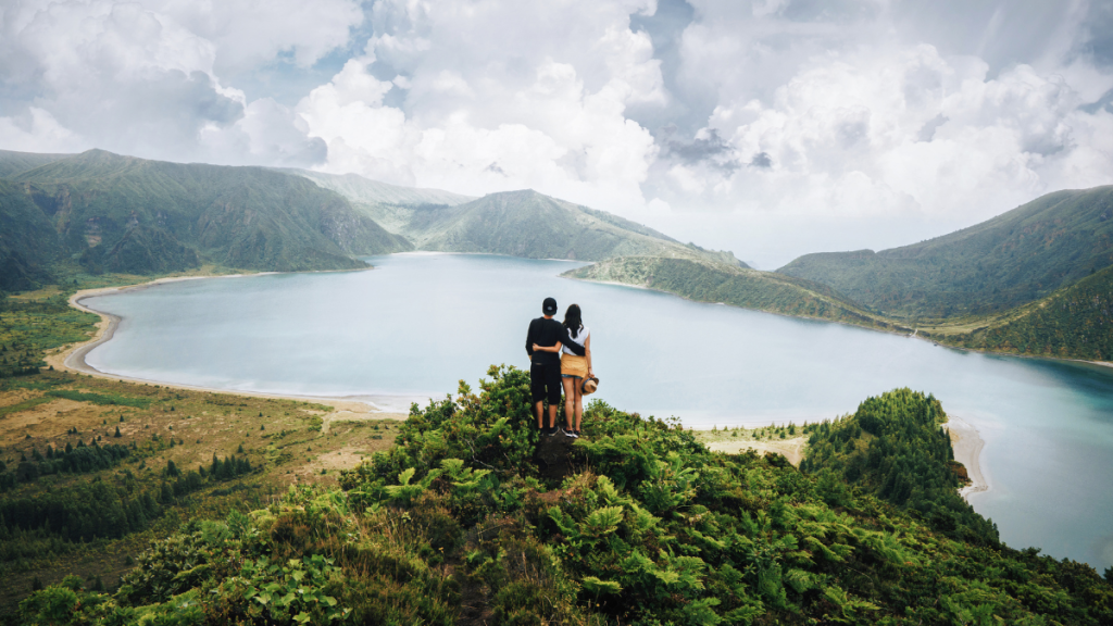 Couple stands on hill overlooking a lake surrounded by mountains.