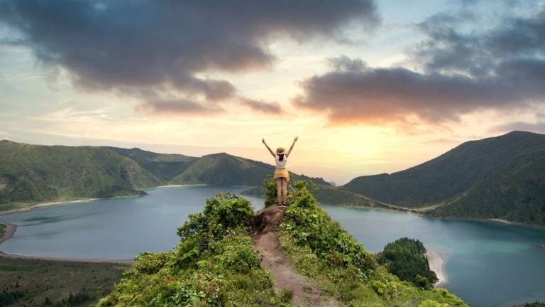 Person standing on a hilltop with arms raised, overlooking a lake and mountains at sunset.