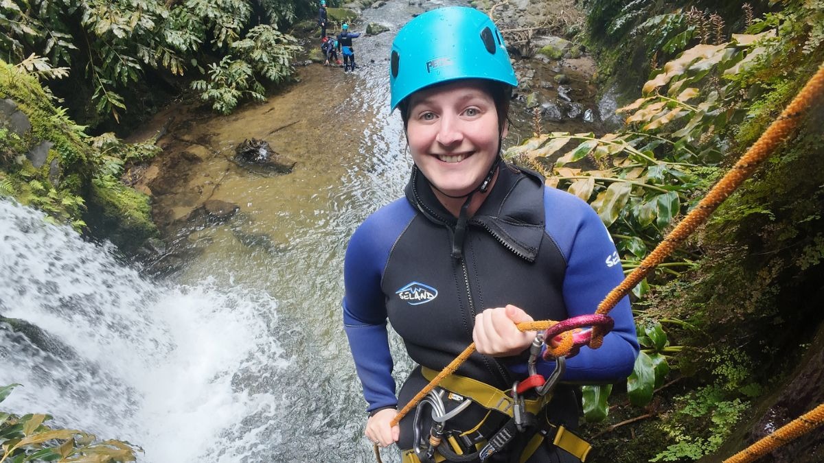 Person in a helmet and wetsuit smiling while rappelling beside a waterfall.