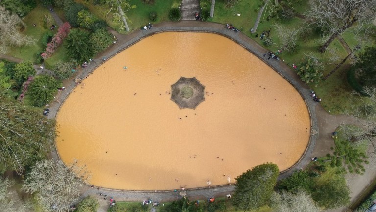 Aerial view of an oval, orange-tinted pond with central fountain, surrounded by trees and pathways.