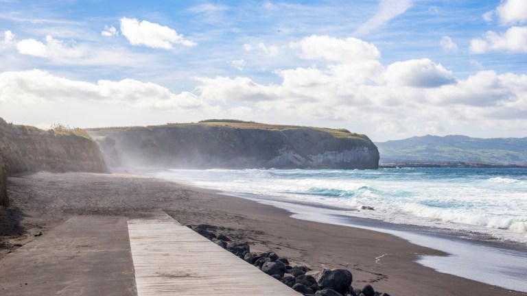 Coastal beach scene with cliffs, waves, and cloudy sky.