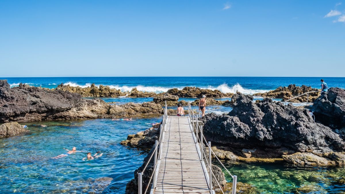 Wooden pier over rocky seashore with people swimming and clear blue sky.