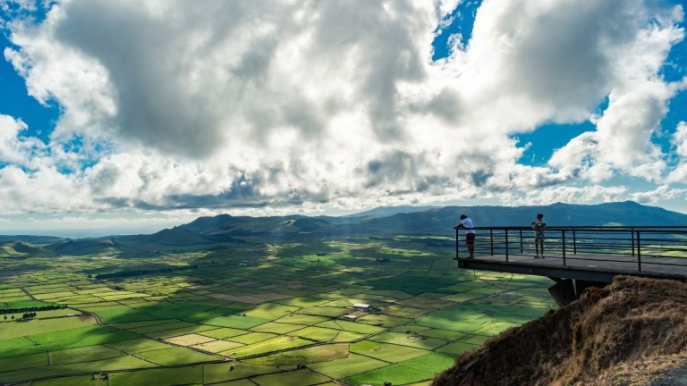 Two people on a viewing platform overlooking a vast, green landscape with scattered clouds.