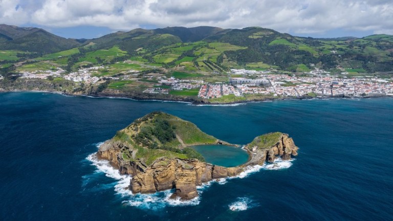 Aerial view of a small island with a lake and cliffs, near a coastal town and green hills.