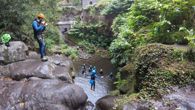 People canyoning in lush, rocky landscape with one person jumping into a river.