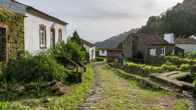 Rural stone path with white and stone houses, greenery, and hills in the background.