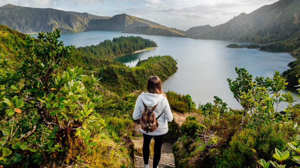 Person with a backpack walks toward a scenic lake surrounded by hills and greenery.