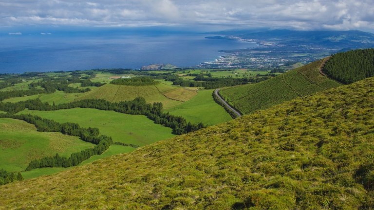 Hilly landscape with green fields, distant coastline, and cloudy sky.