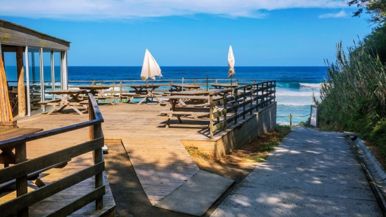 Wooden deck with tables overlooking a beach and ocean under a blue sky.