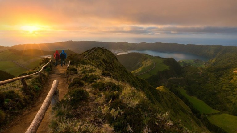 Two hikers on a mountain trail overlooking a sunset view of lakes and greenery.