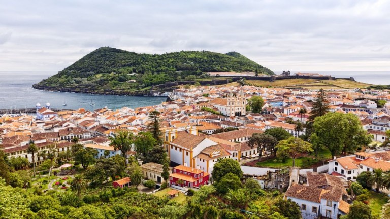 Coastal town with red roofs, lush greenery, and a hilly backdrop near the sea.