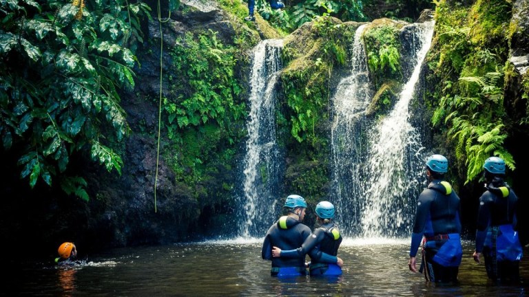 People in wetsuits and helmets near a waterfall in a forest setting.