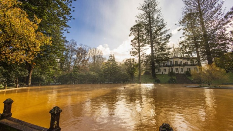 Lake with brown water, surrounded by trees and a house in the background.