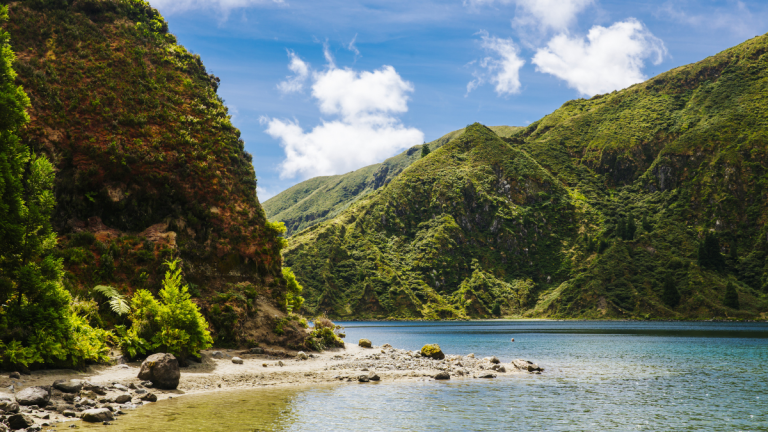 Lush green hills and a rocky shore by a calm lake under a blue sky with clouds.