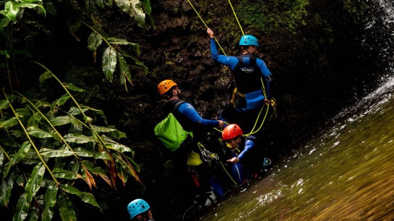 Group of people in helmets and wetsuits climbing a mossy rock near water.
