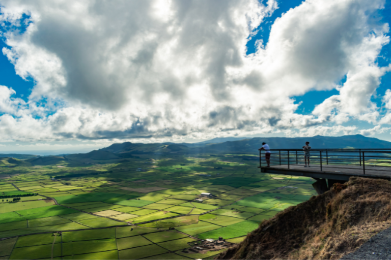 Two people stand on a scenic overlook above green fields under a cloudy sky.
