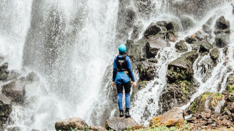 Person in blue gear stands near a waterfall on rocks.