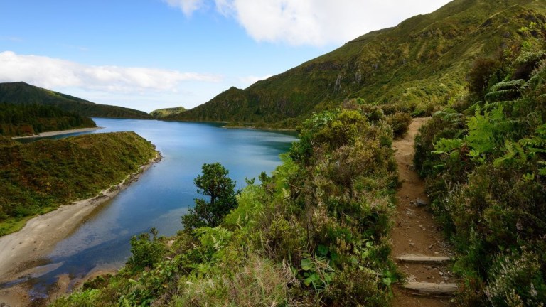 Pathway beside a calm lake with hills and blue sky in the background.