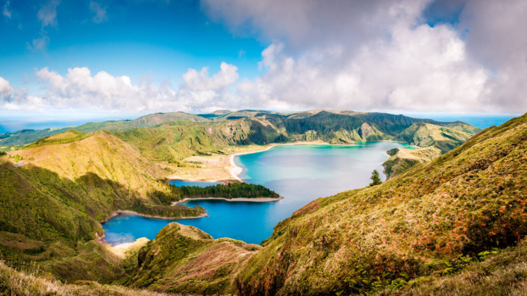 Panoramic view of a lake surrounded by lush green hills under a partly cloudy sky.