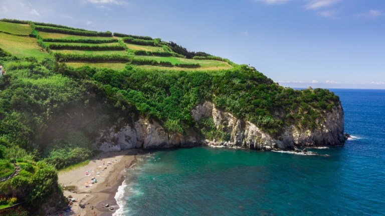Cliffside beach with green fields above and blue ocean below on a sunny day.