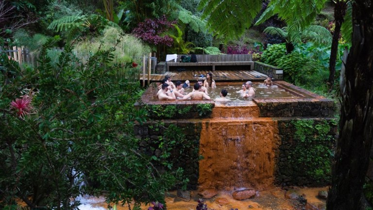 People relax in a natural hot spring pool surrounded by lush greenery and a small waterfall.
