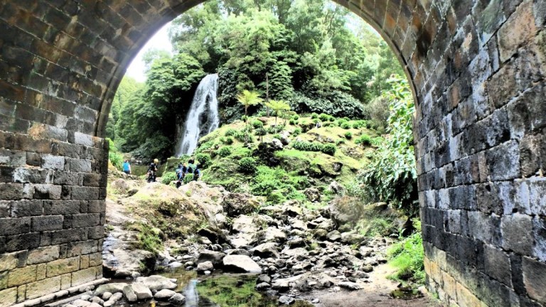 View of a waterfall through a stone arch with lush greenery and rocks.