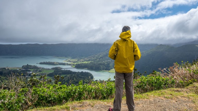 Person in yellow jacket overlooking scenic lake and mountains under a cloudy sky.