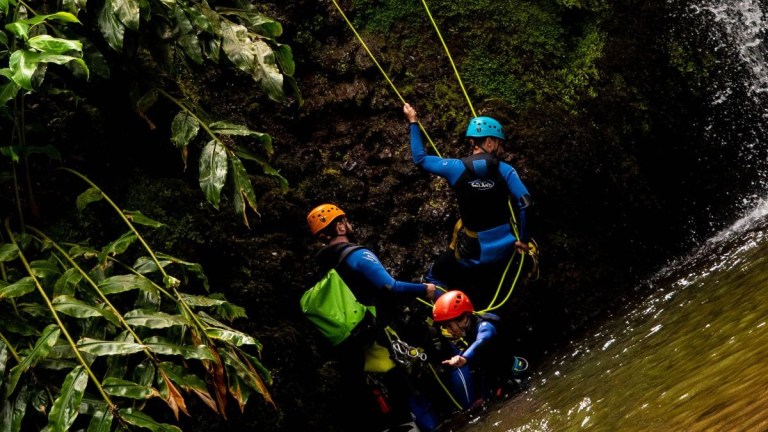Three people in helmets and wetsuits climbing a mossy rock near a waterfall with ropes.