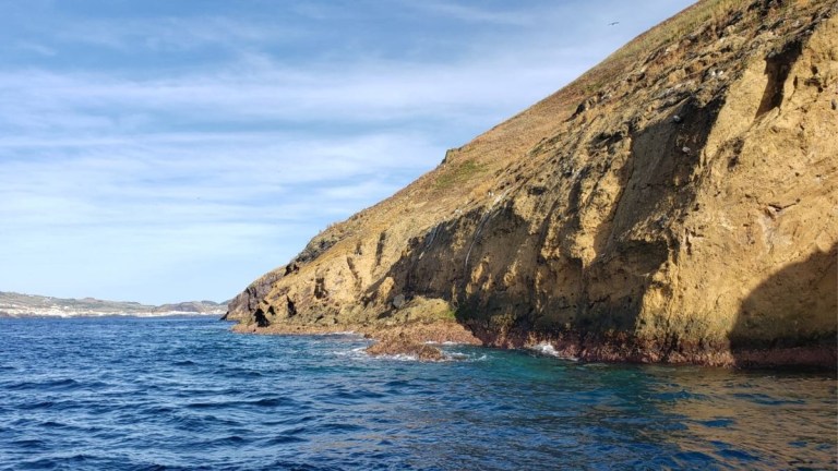 Rocky coastline with clear blue water and a hill under a blue sky.