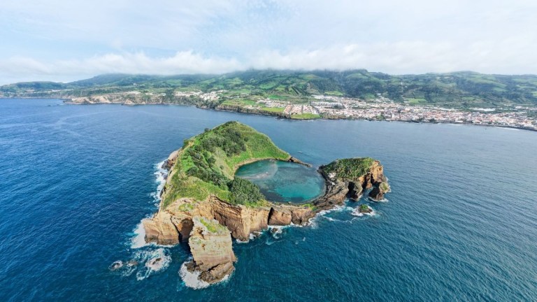 Aerial view of a green island with surrounding blue ocean and distant shoreline city.