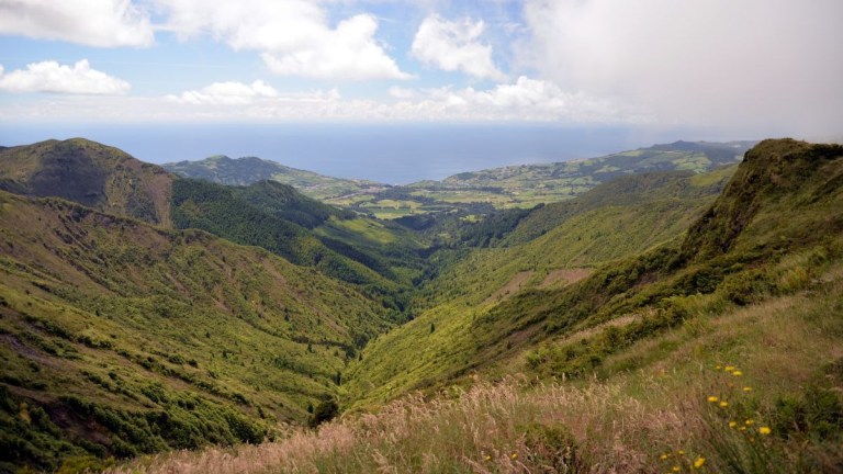 Scenic view of green valley, hills, and distant ocean under a partly cloudy sky.
