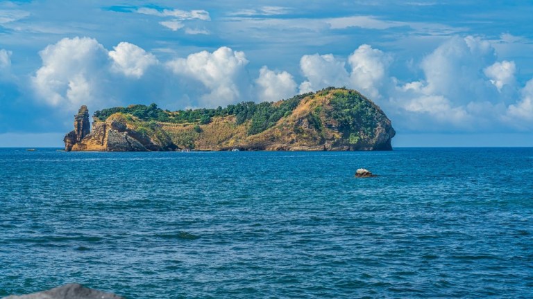 Small green island in blue ocean with cloudy sky.