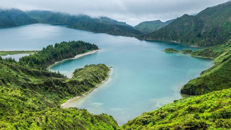 Scenic view of a lake surrounded by lush green hills under a cloudy sky.
