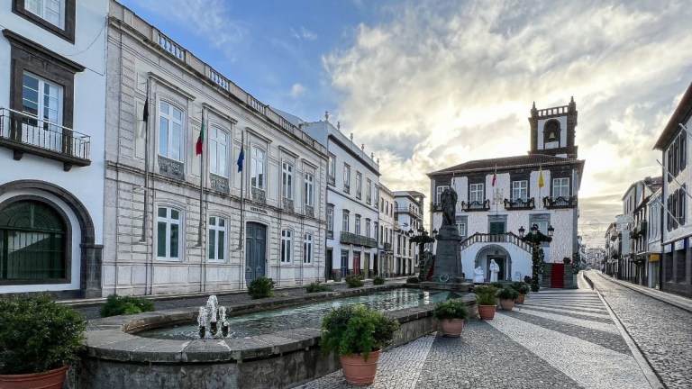Historic European plaza with buildings, fountain, and cloudy sky.
