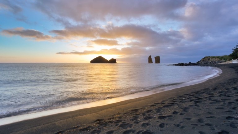 Sunset on a beach with black sand and rock formations in the ocean.