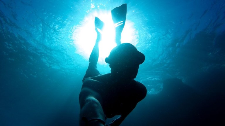 Silhouette of a snorkeler underwater with sunlight overhead.