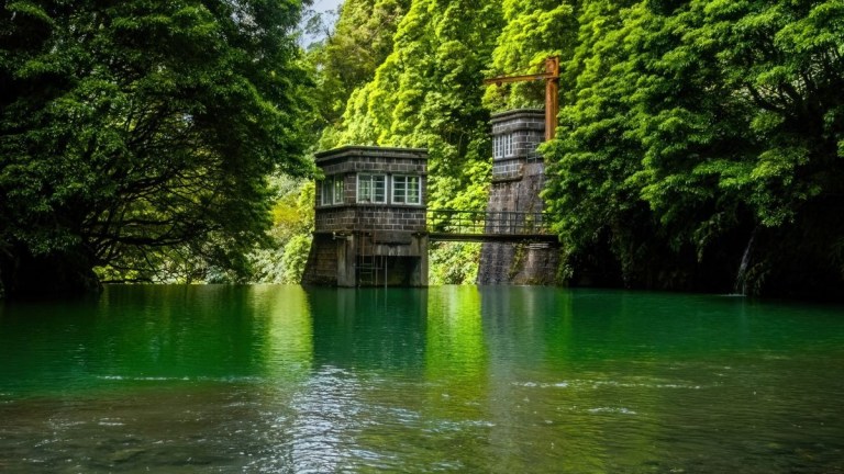 Old wooden structures by a green forested riverbank.