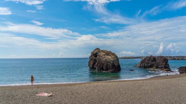 Person walking on sandy beach near large rocks in calm sea under a blue sky.