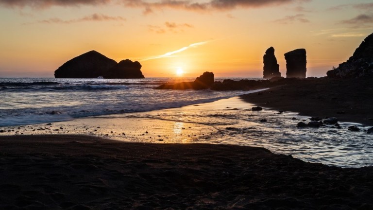 Sunset over ocean with rocky formations and sandy beach.
