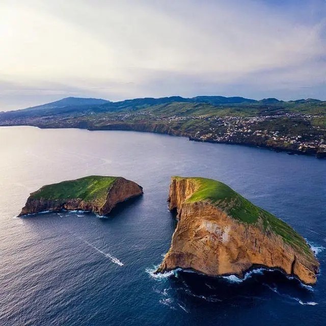 Two rocky, grass-covered islands in a calm sea, with a coastal landscape in the background.