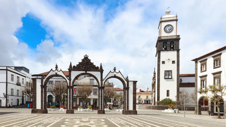 Historic square with arches and clock tower under a cloudy sky.